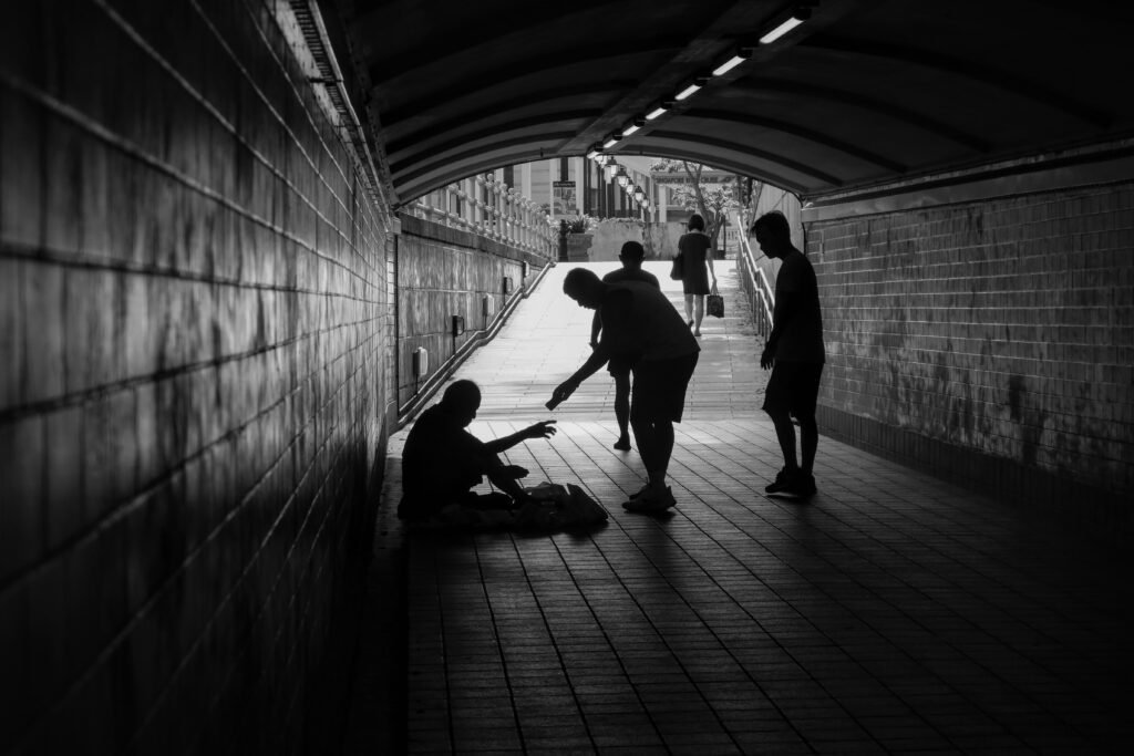 Homem e seu filho oferecendo esmola a um senhor sentado no chão dentro de um túnel, destacando a importância das finanças. A cena simboliza como nossas escolhas financeiras afetam não só nossa vida, mas também a de outras pessoas, lembrando que a educação financeira pode fazer a diferença no caminho que seguimos e na capacidade de ajudar ao próximo.