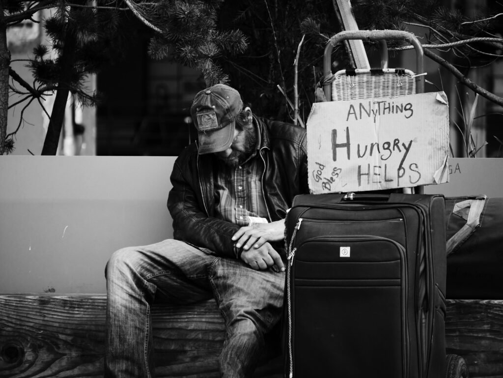 Homem sentado cabisbaixo em um banco de madeira, com uma mochila ao lado e um cartaz pedindo ajuda, escrito 'qualquer coisa, estou com fome' e 'Deus abençoe'. A imagem ilustra a realidade de muitas pessoas que enfrentam dificuldades financeiras e a importância da educação financeira para evitar situações de vulnerabilidade e promover estabilidade e segurança financeira.